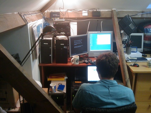 man sitting at computer screen with many servers in loft space. 